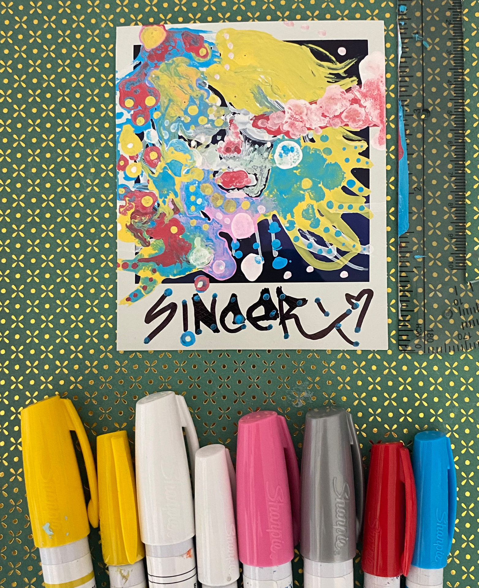 A colorful painted Polaroid in progress, photographed on a patterned surface beside paint markers